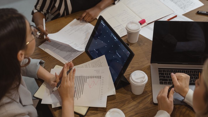Team members discuss project documents and laptops around a meeting table.